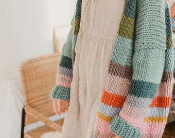 May include: A chunky knit cardigan in a variety of colors, including teal, green, orange, pink, and gray. The oversized sweater has wide sleeves and a striped pattern. The cardigan is worn over a light-colored dress.