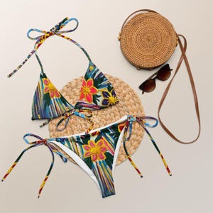 May include: A colorful floral print bikini set with a matching top and bottom. The bikini features a vibrant pattern of flowers in shades of pink, yellow, and blue. A round woven bag and sunglasses are also in the image.