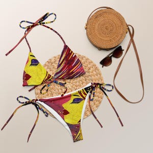 May include: A two-piece bikini set with a vibrant face design in yellow, red, and purple. The bikini top and bottom feature tie closures. A round woven bag, sunglasses, and a straw mat complete the beach-themed ensemble.