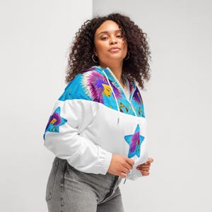 May include: A white and blue cropped windbreaker jacket featuring a vibrant graphic print of a stylized figure with purple hair inside a blue star, accented with small gold Libra scale symbols across the upper chest and sleeves, with a hood.