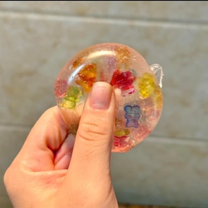 Op de afbeelding: Een heldere, ronde hars papeterie met kleurrijke gummyberen. De gummyberen zijn rood, geel, groen en blauw. De papeterie wordt in een hand gehouden.