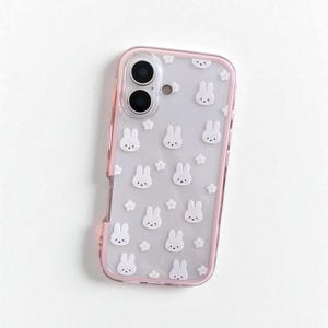 以下が含まれることがあります： ピンクの縁取りと白いウサギの顔と小さな白い花の模様が特徴の透明な携帯電話ケース。ケースは携帯電話を保護し、キュートなデザインです。