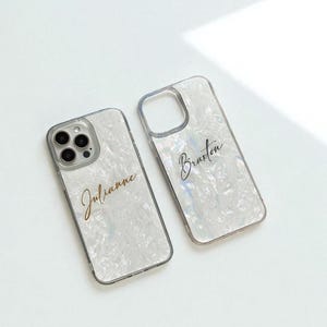 以下が含まれることがあります： 真珠光沢のデザインの透明な携帯電話ケースが2つ。1つのケースには「Julianne」という名前が金色の筆記体で、もう1つには「Braxton」という名前が金色の筆記体で書かれています。ケースは銀色の縁取りで、白い背景にあります。