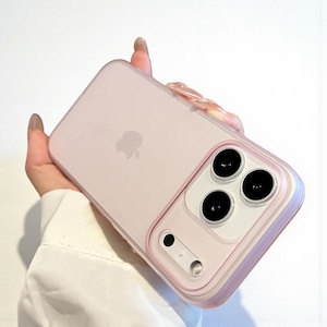 May include: A pale pink iPhone case with a transparent edge. The case features a camera aperture with three black lenses and a flash. The Apple logo is visible on the back. The case is held in a hand.