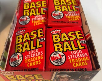 May include: A box of 1982 Fleer Baseball trading cards with a red, white, and blue color scheme. The box features a sticker of a baseball player and the text "BASEBALL 1982 TEAM LOGO STICKER TRADING CARDS".