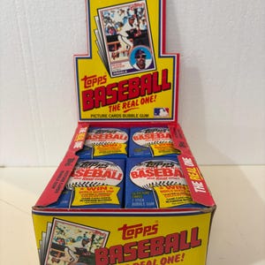 May include: A box of Topps Baseball cards with a red, white, and blue design. The box features a baseball and the text "Topps Baseball The Real One!"  The box contains 15 picture cards and 1 stick of bubble gum.