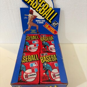 May include: A blue cardboard baseball card display box with the word "BASEBALL" in yellow. The box contains multiple red packs of baseball cards, each with a baseball graphic and the text "18 CARDS". The display also features a baseball player illustration.