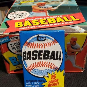 May include: A blue baseball card pack with a baseball graphic and the word "BASEBALL" in large letters. The pack is in front of a box of baseball cards. The box has the words "MAJOR LEAGUE BASEBALL" and "PICTURE CARDS BUBBLE GUM".