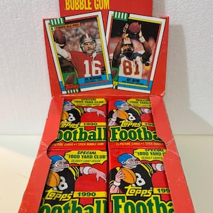 May include: A vintage Topps Football picture card box with packs of cards and bubble gum. The box is red with green and yellow text, featuring images of football players and the Topps logo. The box includes packs of 15 picture cards and 1 stick of bubble gum.