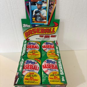 May include: A green and white Topps Baseball card display box filled with baseball card packs. The box features a Ken Griffey Jr. baseball card and the text "BASEBALL THE REAL ONE!" The display also includes a 50¢ price tag.