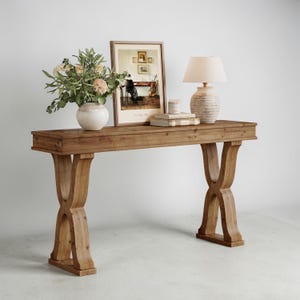 May include: A rectangular wooden console table with X-shaped legs, topped with a white ceramic vase containing green foliage, a framed print of an interior scene, two stacked books, a small candle, and a beige table lamp with a textured base.