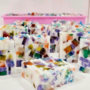 Puede incluir: Barras de jabón hechas a mano con un diseño de mosaico. Los jabones son blancos con piezas coloridas de forma irregular en tonos morado, naranja, verde y azul. En el fondo se ve un molde rectangular rosa.
