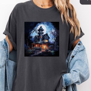 May include: Dark gray t-shirt featuring a spooky haunted house graphic under a full moon. The house is lit up with warm lights, surrounded by bare trees, and set against a dark blue night sky. The word "Pepper" is in a black box in the upper right corner.
