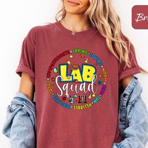 Pode incluir: Uma camiseta cor de tijolo com as palavras "LAB Squad" em um design circular colorido. O design inclui ícones relacionados ao laboratório e palavras como "caring" e "reliable". A camiseta é usada com uma jaqueta jeans.