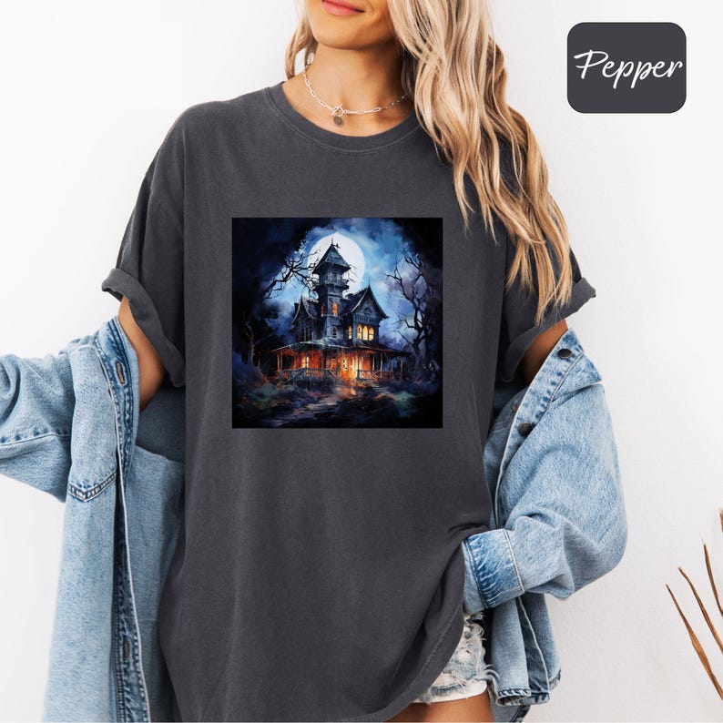 May include: Dark gray t-shirt featuring a spooky haunted house graphic under a full moon. The house is lit up with warm lights, surrounded by bare trees, and set against a dark blue night sky. The word "Pepper" is in a black box in the upper right corner.
