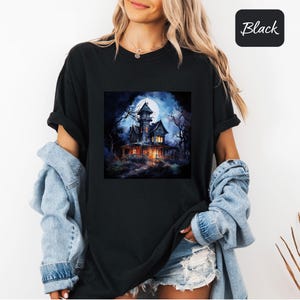 May include: Black t-shirt featuring a spooky haunted house illustration under a full moon. The house is lit up with warm lights, surrounded by dark trees. The word "Black" is in a black square in the upper right corner.