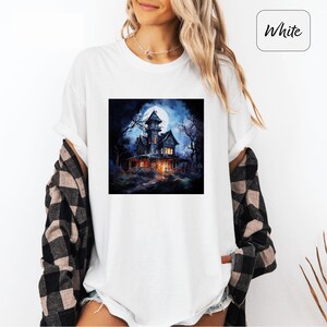 May include: White t-shirt featuring a square graphic of a spooky haunted house under a full moon. The house is dark with lit windows, surrounded by trees. The word "White" is in a white box in the upper right corner.