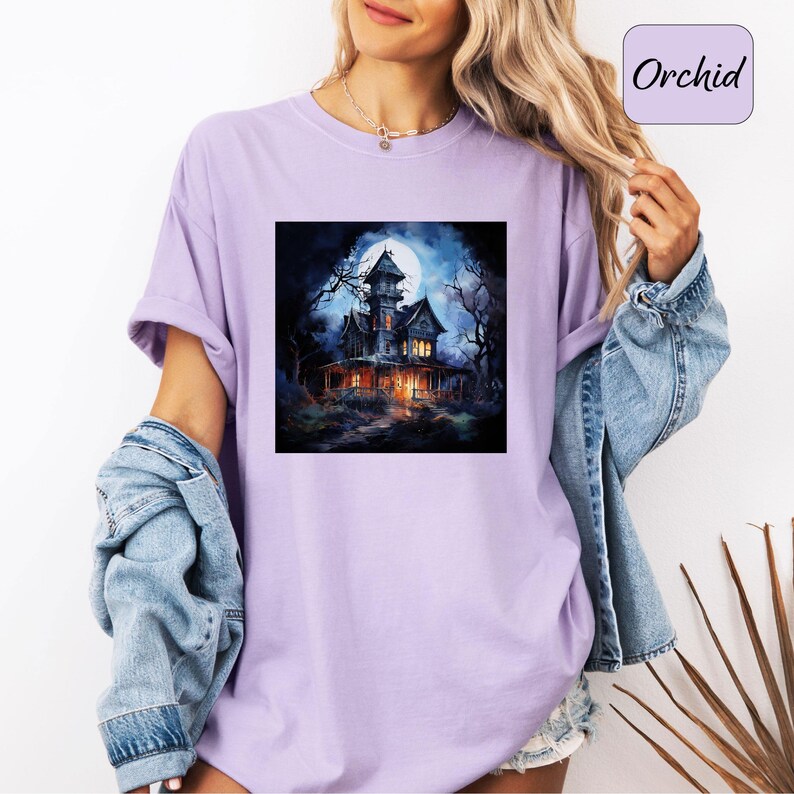 May include: A light purple t-shirt featuring a square graphic of a spooky haunted house under a full moon. The house is dark with lit windows, surrounded by bare trees. The word "Orchid" is in a purple box.
