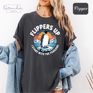 Könnte beinhalten: Ein dunkelgraues T-Shirt mit einem Aufdruck eines Pinguins, der auf einem Eisberg steht. Der Aufdruck enthält den Text "Flippers Up I Stand With The Penguins".
