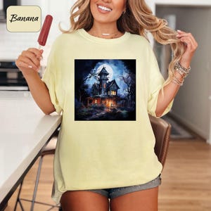 May include: A light yellow t-shirt with a square graphic of a haunted house under a full moon. The house is dark with lit windows, surrounded by bare trees. The word "Banana" is in a yellow box in the upper left corner.