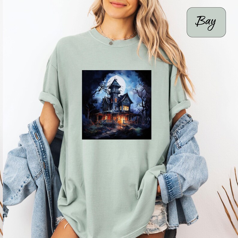 May include: A light green t-shirt featuring a square graphic of a spooky haunted house under a full moon. The house is dark with lit windows, surrounded by bare trees. The word "Bay" is in a light green rectangle in the upper right corner.