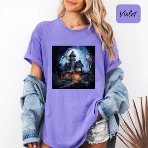 May include: A violet-colored t-shirt with a square graphic of a spooky haunted house under a full moon. The house is lit up with warm lights, surrounded by bare trees, and set against a dark, starry sky. The word "Violet" is in a purple square.
