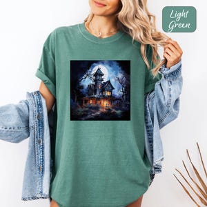 May include: Light green t-shirt featuring a square graphic of a spooky haunted house under a full moon. The house is dark with lit windows, surrounded by bare trees. The words "Light Green" are in a box in the upper right corner.