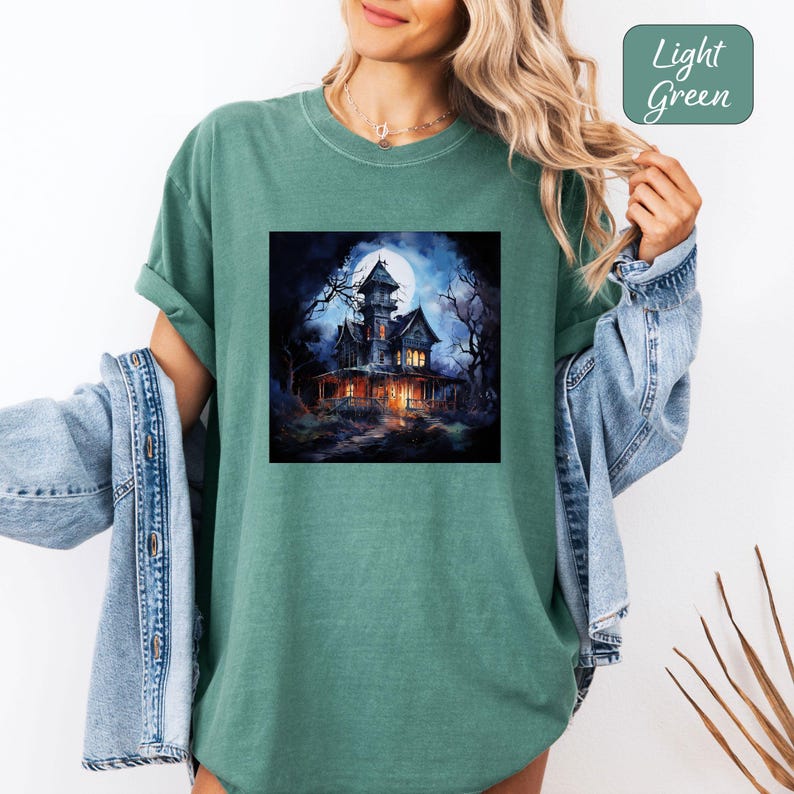 May include: Light green t-shirt featuring a square graphic of a spooky haunted house under a full moon. The house is dark with lit windows, surrounded by bare trees. The words "Light Green" are in a box in the upper right corner.