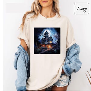 May include: Ivory-colored t-shirt featuring a square graphic of a spooky haunted house under a full moon. The house is dark with lit windows, surrounded by bare trees, creating a Halloween or horror theme.