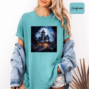 May include: Seafoam green t-shirt featuring a square graphic of a spooky haunted house under a full moon. The house is dark with lit windows, surrounded by bare trees. The shirt has a relaxed fit and rolled-up sleeves.