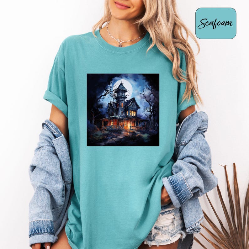 May include: Seafoam green t-shirt featuring a square graphic of a spooky haunted house under a full moon. The house is dark with lit windows, surrounded by bare trees. The shirt has a relaxed fit and rolled-up sleeves.