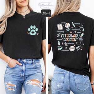 Op de afbeelding: Zwart T-shirt met een pootprint en een kruis op de voorkant. De achterkant van het shirt heeft een ontwerp met veterinaire gereedschappen en de tekst "Veterinary Assistant".