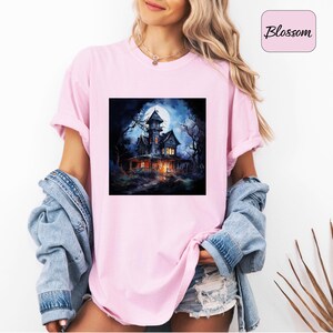 May include: A light pink t-shirt featuring a square graphic of a spooky haunted house under a full moon. The house is dark with lit windows, surrounded by bare trees. The word "Blossom" is in a pink box.
