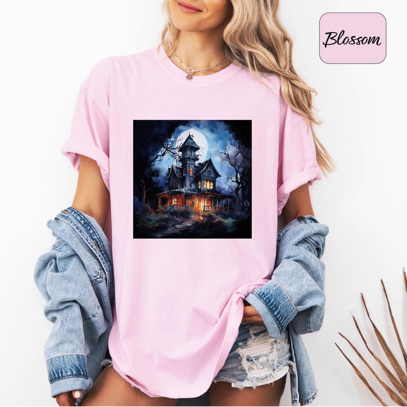May include: A light pink t-shirt featuring a square graphic of a spooky haunted house under a full moon. The house is dark with lit windows, surrounded by bare trees. The word "Blossom" is in a pink box.