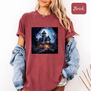 May include: Brick-colored t-shirt featuring a square graphic of a spooky haunted house under a full moon. The house is lit with warm lights, surrounded by dark trees, and set against a dark blue night sky. The word "Brick" is in a red square.