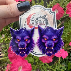 May include: A pair of purple wolf earrings with orange eyes and bared teeth. The earrings are held in front of a sign that reads "Raven Hill". The background features pink and red flowers.
