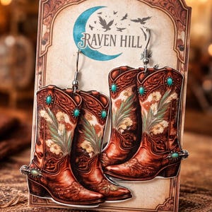 May include: A pair of earrings shaped like cowboy boots, with intricate floral designs and turquoise accents. The earrings are displayed on a card with the text "Raven Hill" and a crescent moon design. The boots are a reddish-brown color.