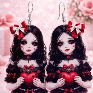 May include: A pair of dangle earrings featuring a gothic doll design. Each earring depicts a doll with black hair, a red bow, and a red heart. The doll is wearing a black dress with a red bow. The earrings have silver hooks.