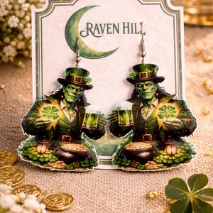 May include: Raven Hill earrings featuring a green-skinned figure in a green hat and jacket, holding a beer and a glowing four-leaf clover. The earrings are set against a backdrop with the Raven Hill logo and gold coins.