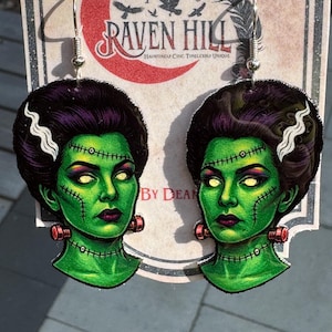 May include: A pair of dangle earrings featuring a green-skinned monster face with black hair, white streaks, and red neck bolts. The earrings are attached to silver hooks. The background features the text "Raven Hill" and "Hauntingly Chic. Timelessly Unique."