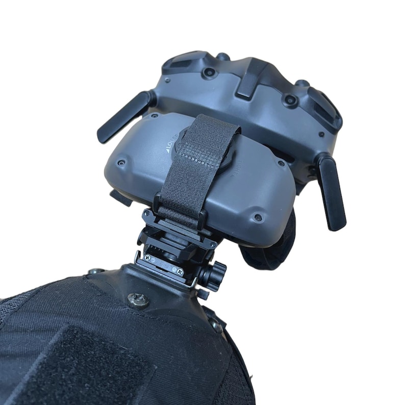 May include: A gray tactical headset with a black strap and mounting hardware. The headset is attached to a black fabric surface, likely a helmet or vest. The image is set against a white background.