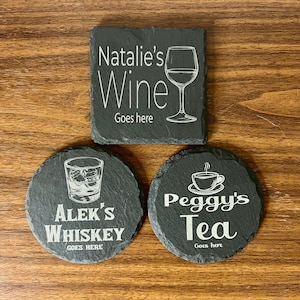 May include: Three black slate coasters with white engraved text and images. The first coaster says "Natalie's Wine Goes here" and has a wine glass image. The second coaster says "Alek's Whiskey Goes here" and has a whiskey glass image. The third coaster says "Peggy's Tea Goes here" and has a teacup image.