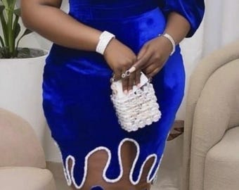 Könnte beinhalten: Ein königsblaues Samtkleid mit einzigartigem Design. Das One-Shoulder-Kleid hat weiße Verzierungen und einen transparenten Einsatz mit blauen Akzenten. Eine kleine, weiße Perlenhandtasche wird gehalten.