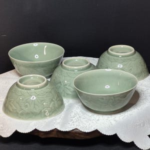 May include: Five celadon ceramic bowls of varying sizes are arranged on a white, scalloped-edge cloth. The bowls have a glossy finish and decorative floral patterns. The background is a dark, solid colour.