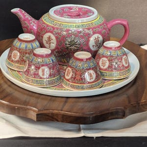Może przedstawiać: Różowy ceramiczny zestaw do herbaty z imbrykiem i pięcioma pasującymi filiżankami. Zestaw posiada skomplikowane wzory kwiatowe i geometryczne w kolorze czerwonym, żółtym i białym. Zestaw do herbaty jest prezentowany na białej tacy i drewnianej podstawie.