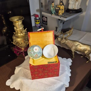 Peut inclure: Une boîte rouge à motifs avec un intérieur jaune contient deux bols. L'un est blanc, l'autre est décoré d'un motif floral vert, bleu et rouge. La boîte repose sur un tissu en dentelle blanche.