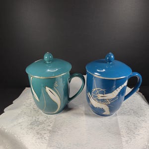 May include: Two ceramic mugs with lids, one teal and one blue, each with a gold rim and handle. The teal mug features white leaf designs, while the blue mug has a white abstract design. A small figurine is in the background.
