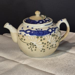 Può includere: Una teiera in ceramica con corpo arrotondato e coperchio. La teiera è principalmente beige con disegni dipinti in blu e bianco. Il manico e il beccuccio sono bianchi. Il coperchio ha un piccolo pomello ed è decorato con accenti verdi. La teiera è su una superficie bianca.