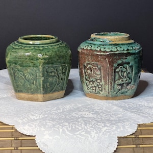 Pode incluir: Dois potes de cerâmica antigos com desenhos em relevo. Um tem formato hexagonal com esmalte verde, o outro é redondo com esmalte marrom e verde-azulado. Ambos têm uma aparência desgastada, sugerindo idade e uso. Estão sobre um pano branco.