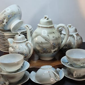 Può includere: Un servizio da tè in porcellana vintage con un motivo a gru e floreale. Il set include una teiera, zuccheriera, lattiera, tazze e piattini. Il set da tè è bianco con un disegno dipinto a mano in blu, grigio e oro.